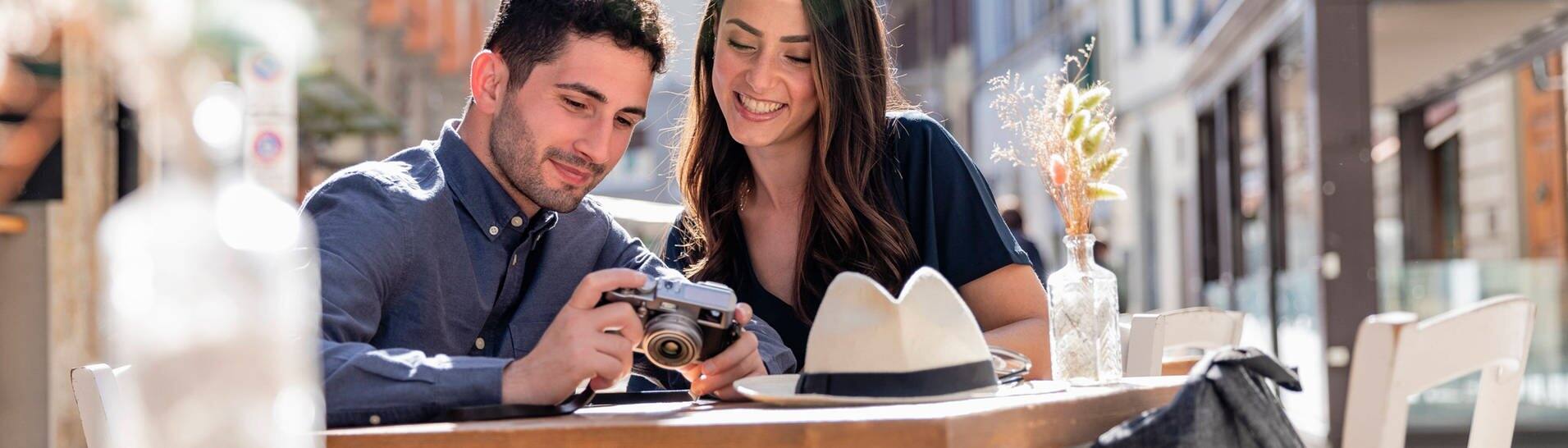 Ein Pärchen Touristen beim Städtetrip. Sie schauen sich Fotos auf einer Kamera an und sitzen in einem Café einer Stadt . (Foto: IMAGO Westend61)