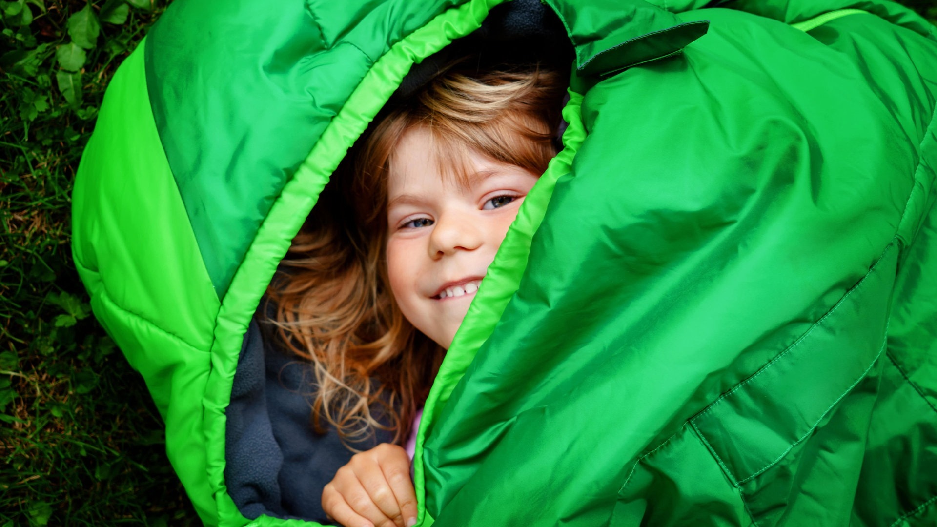 Foto: Adobe Stock, Irina Schmidt Kleines Kind liegt grinsend auf dem Boden im knallgrünen Schlafsack (Foto: Adobe Stock, Irina Schmidt)