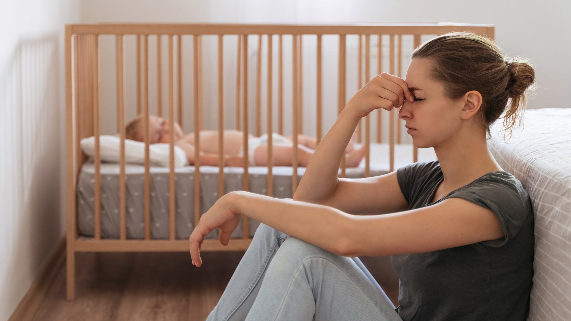 Frau mit Wochenbettdepression sitzt am Boden neben ihrem Baby (Foto: Adobe Stock, Arsenii)
