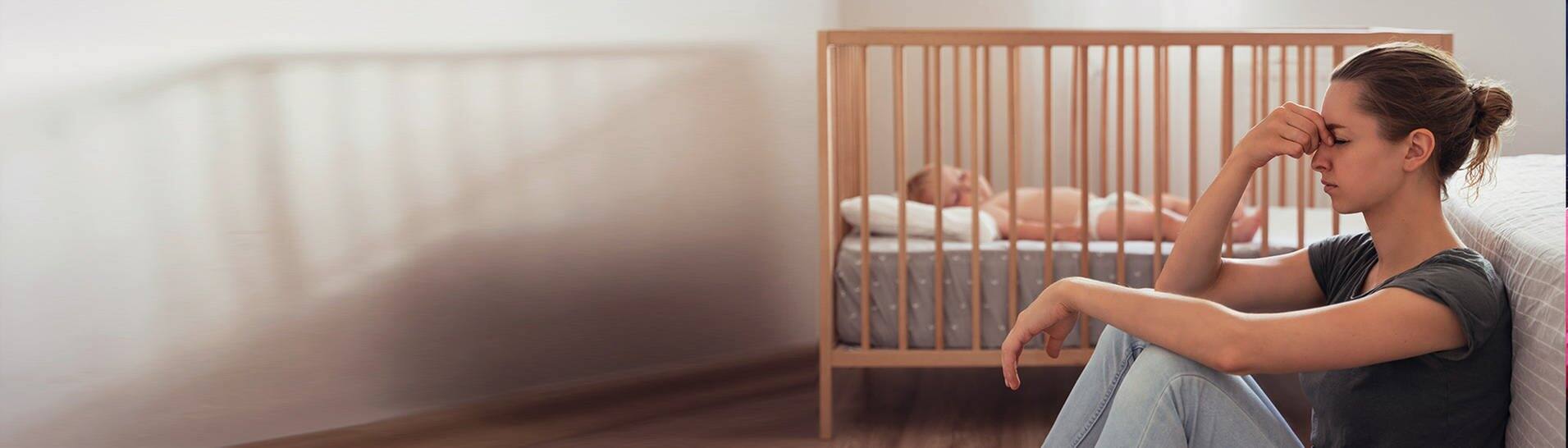 Frau mit Wochenbettdepression sitzt am Boden neben ihrem Baby (Foto: Adobe Stock, Arsenii)