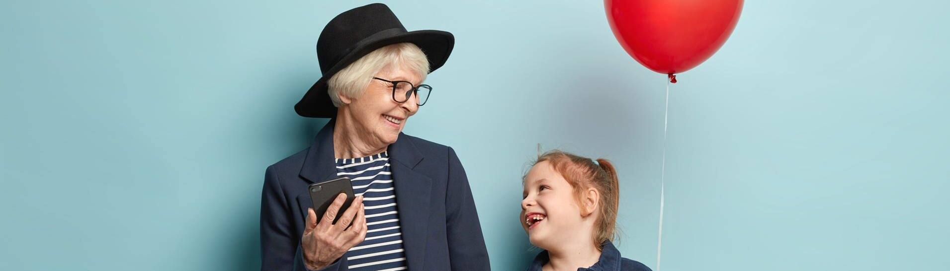 Eine ältere Frau hält ein Handy in der einen und ihre Enketochter in der anderen Hand, die wiederum einen Luftballon in der Hand hält (Foto: Adobe Stock, Wayhome Studio)