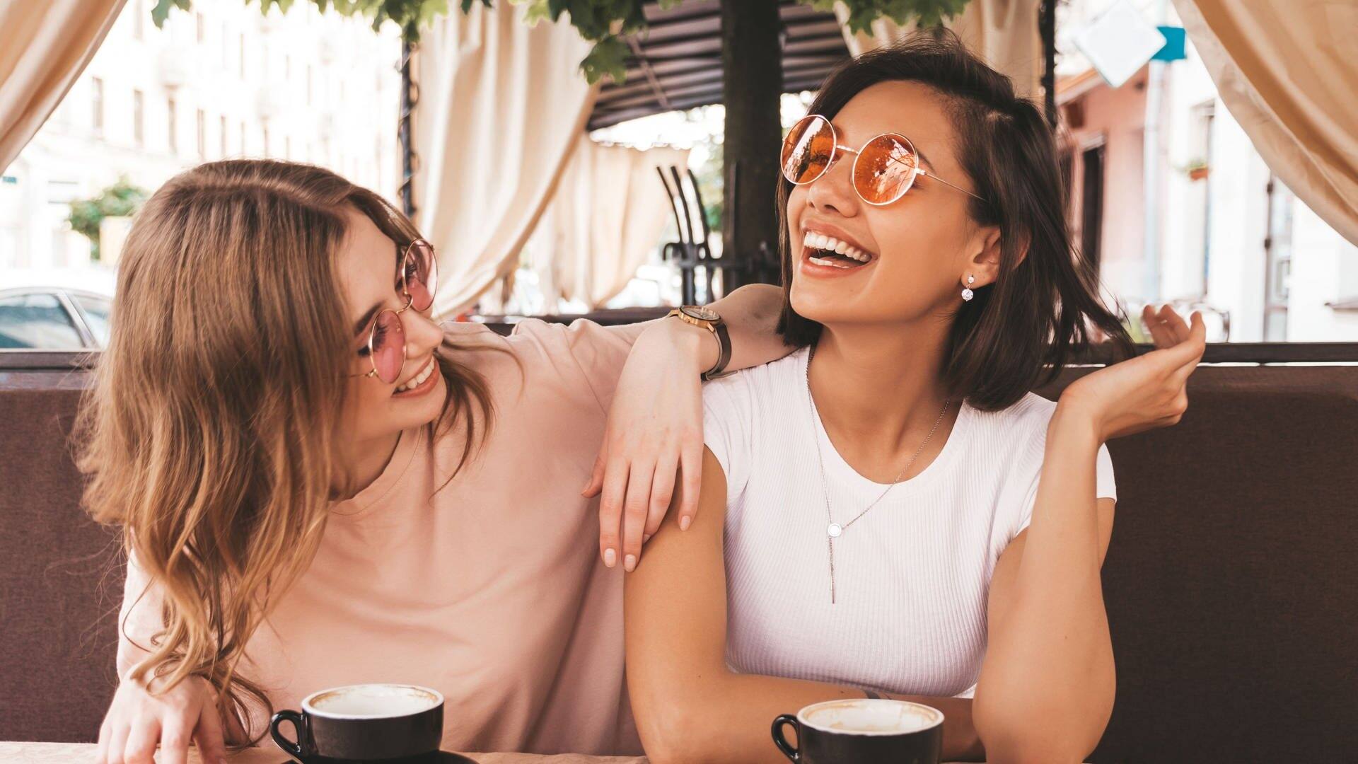 Zwei Freundinnen sitzen in einem Café, vor ihnen auf dem Tisch stehen die Getränke. Sie lachen gemeinsam, vielleicht über die Storys aus Partnerschaft und Liebe, die sie einander erzählen. (Foto: Adobe Stock, halayalex)
