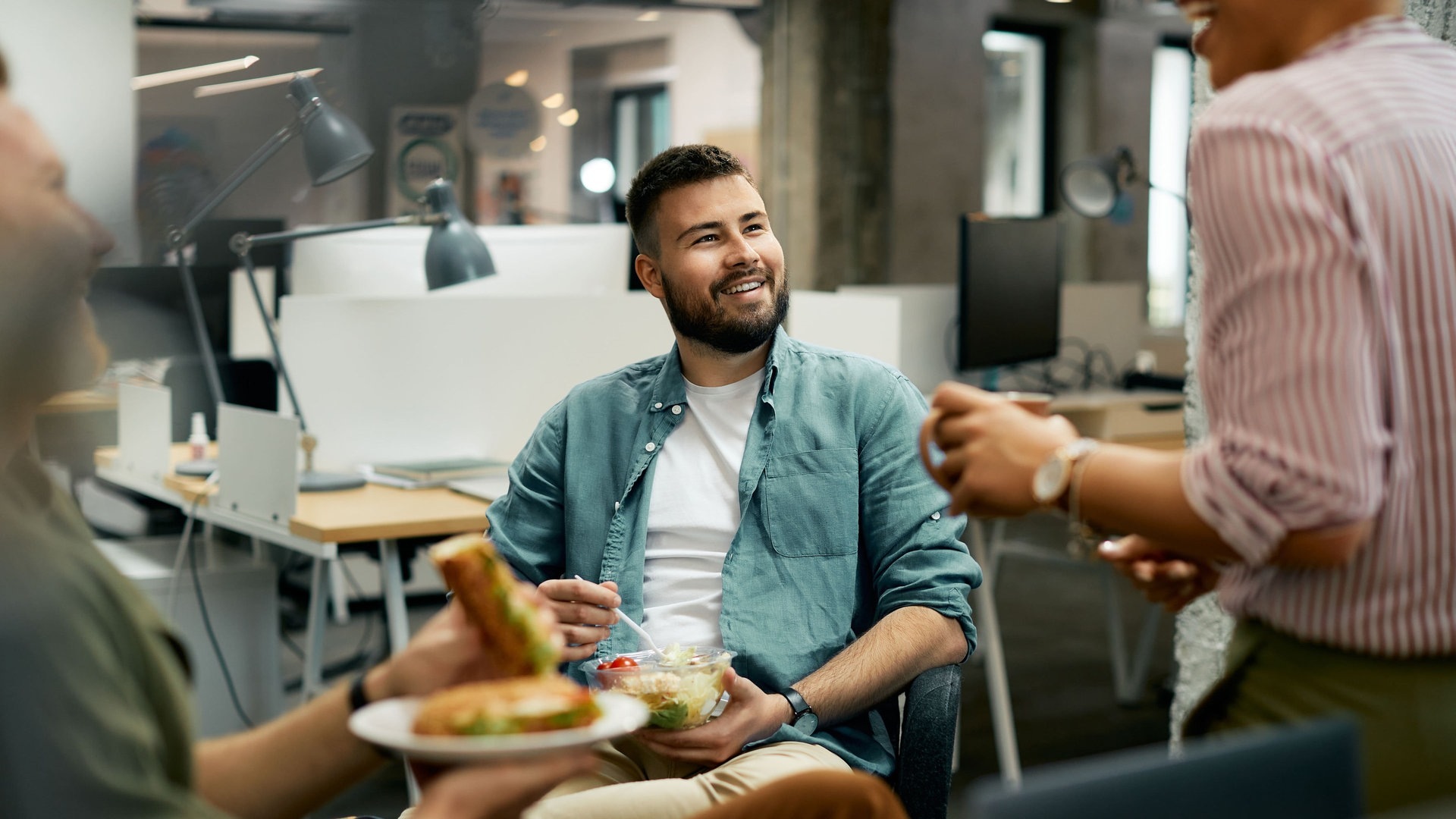 Foto: Adobe Stock, Drazen Drei Leute, die wohl miteinander arbeiten, machen gemeinsam eine Pause, so wie es im Gesetz verordnet ist. Sie essen etwas im Büro und lachen miteinander. (Foto: Adobe Stock, Drazen)