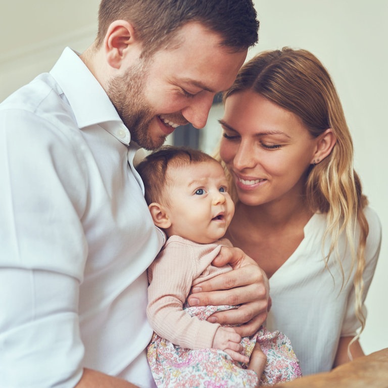 Junge Eltern mit ihrem Baby. Der Vater hält lachend das Baby auf dem Arm, die Mutter lächelt das Kind ebenfalls an. (Foto: Adobe Stock, Flamingo Images)