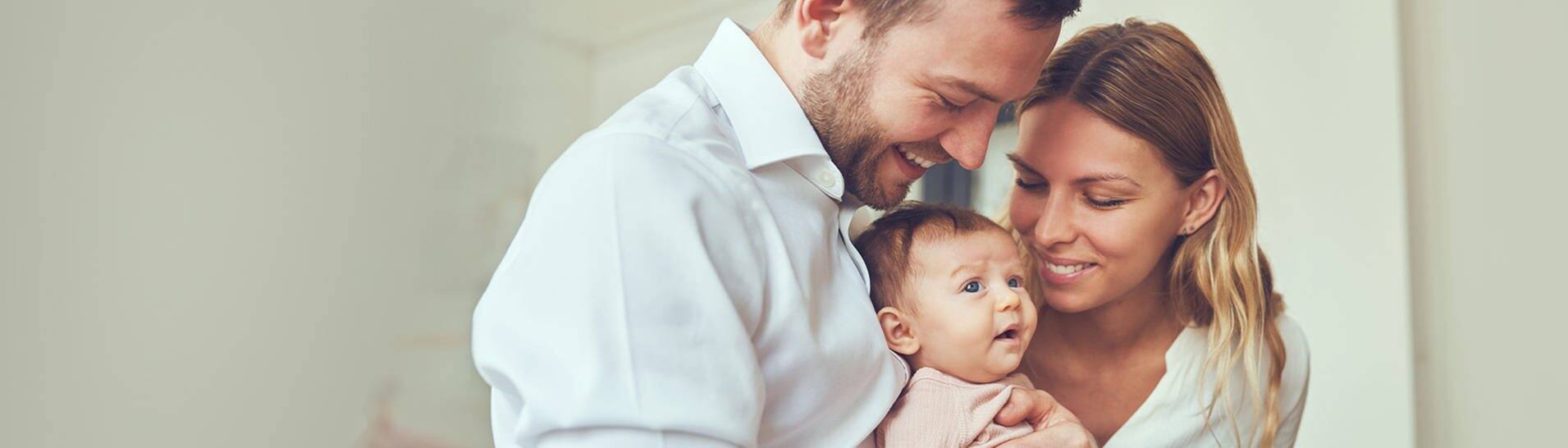 Junge Eltern mit ihrem Baby. Der Vater hält lachend das Baby auf dem Arm, die Mutter lächelt das Kind ebenfalls an. (Foto: Adobe Stock, Flamingo Images)