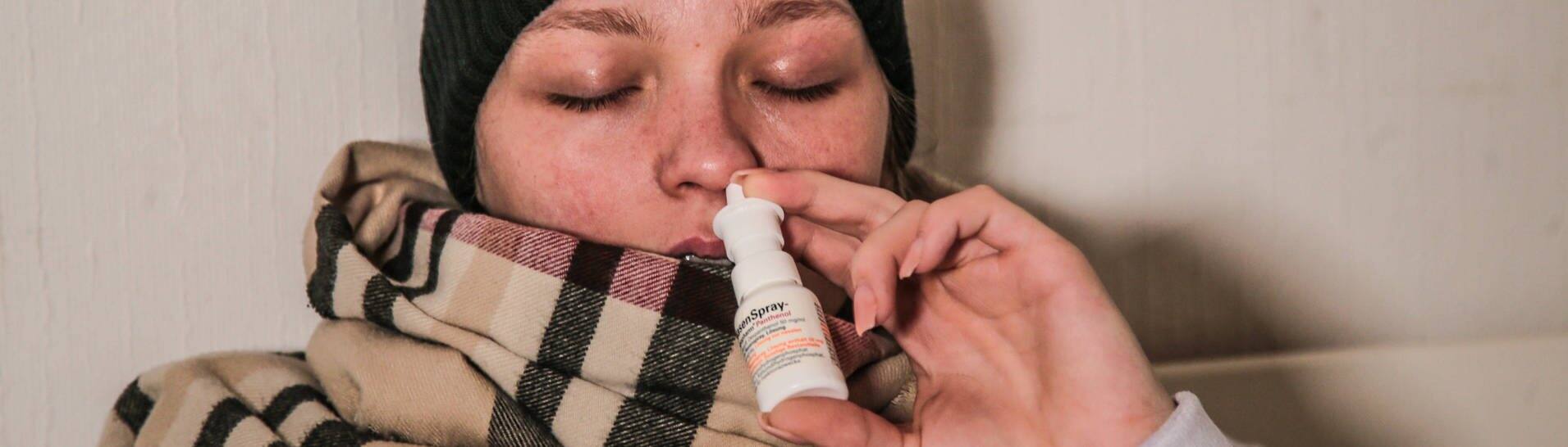 Kranke Frau mit Schal und schwarzer Mütze nutzt ein Nasenspray (Foto: IMAGO, impfung-gegen-grippe)