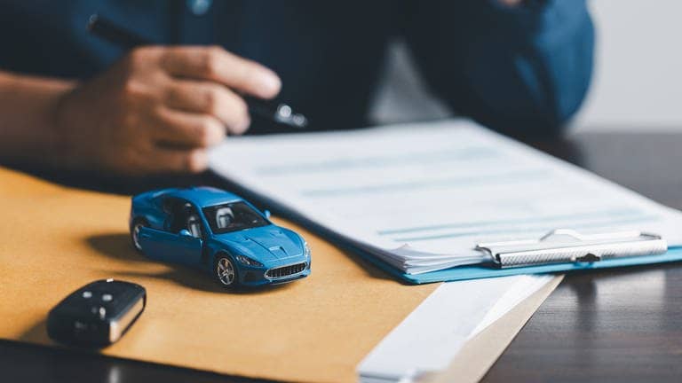 Blaues Miniaturauto liegt auf einem Tisch. Daneben ein richtiger Autoschlüssel, im Hintergrund eine hand mit einem Stift. (Foto: Adobe Stock, JD8)