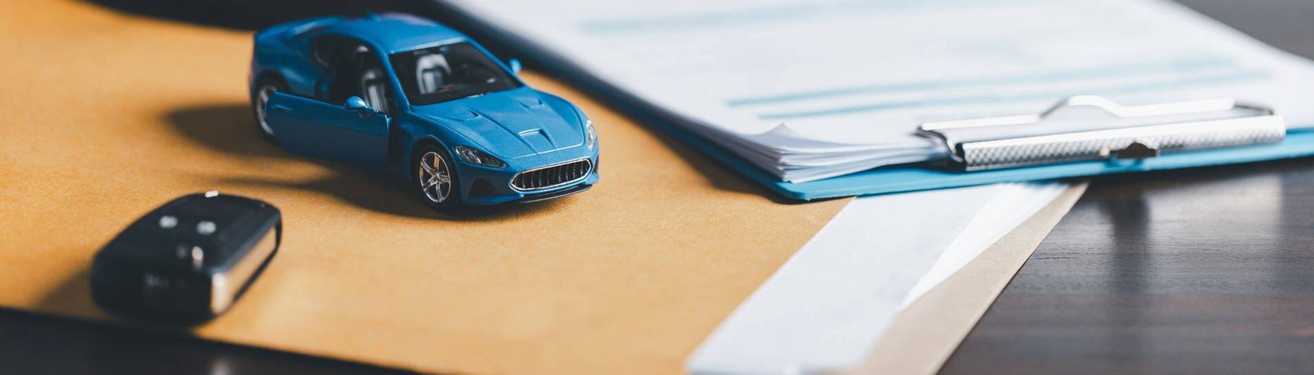 Blaues Miniaturauto liegt auf einem Tisch. Daneben ein richtiger Autoschlüssel, im Hintergrund eine hand mit einem Stift. (Foto: Adobe Stock, JD8)