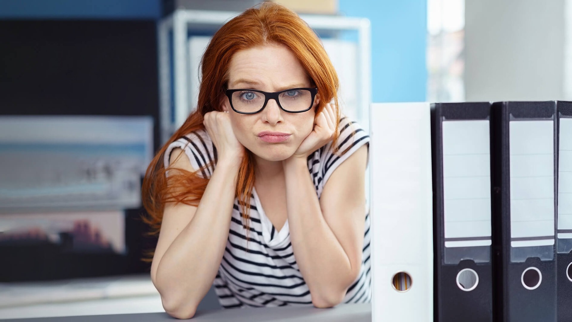 Junge Arbeitnehmerin sitzt mit frustriertem, fragendem Gesichtsausdruck bei der Arbeit und denkt über die Rechte und Konsequenzen von Arbeitsverweigerung nach. Neben ihr sind Aktenordner auf dem Tisch. (Foto: Adobe Stock, contrastwerkstatt)
