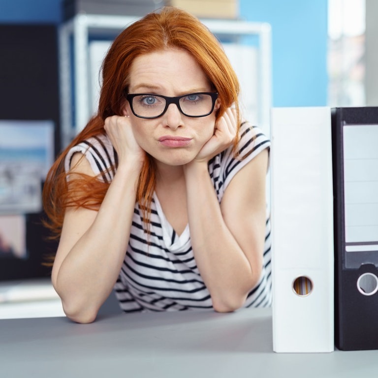 Junge Arbeitnehmerin sitzt mit frustriertem, fragendem Gesichtsausdruck bei der Arbeit und denkt über die Rechte und Konsequenzen von Arbeitsverweigerung nach. Neben ihr sind Aktenordner auf dem Tisch. (Foto: Adobe Stock, contrastwerkstatt)