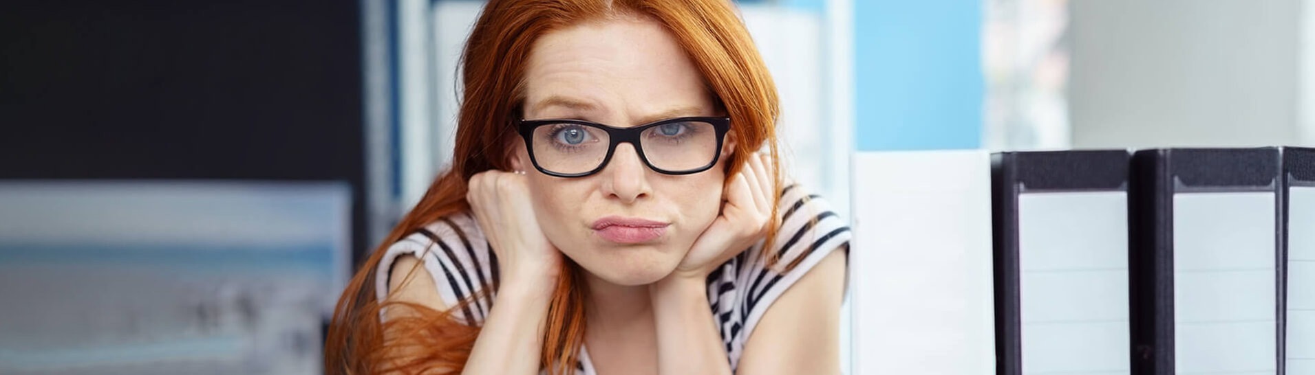 Junge Arbeitnehmerin sitzt mit frustriertem, fragendem Gesichtsausdruck bei der Arbeit und denkt über die Rechte und Konsequenzen von Arbeitsverweigerung nach. Neben ihr sind Aktenordner auf dem Tisch. (Foto: Adobe Stock, contrastwerkstatt)