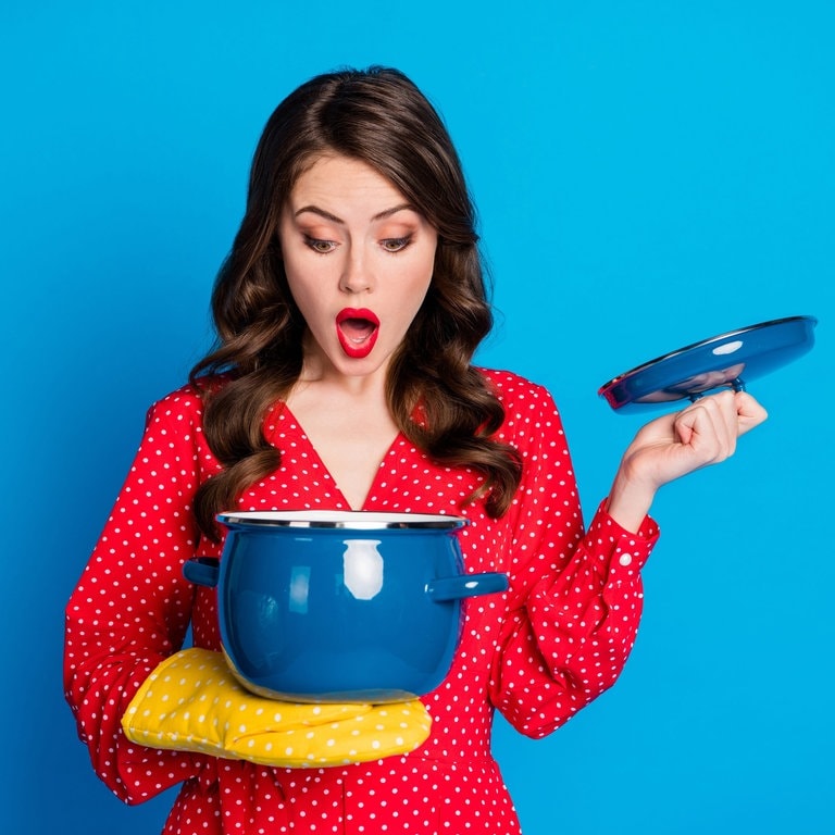 Junge Frau mit Koch-Handschuh schaut verwundert in einen Kochtopf. Da drin ist bestimmt Hühnersuppe gegen Erkältung. (Foto: Adobe Stock, deagreez)