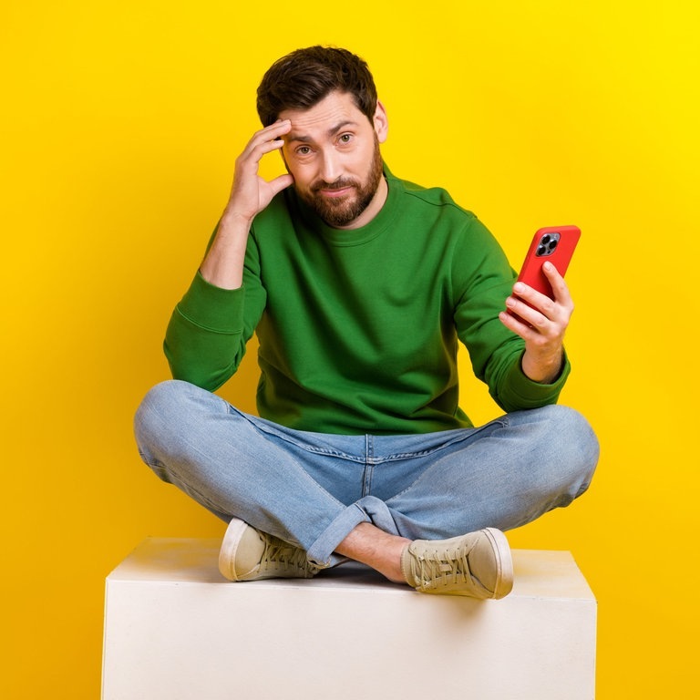 Junger Mann sitzt im Schneidersitz und mit dem Handy in der Hand auf einem Hocker. Er häkt denkend die Hand an den Kopf. Auf seinem Smartphone sieht er bestimmt gerade die überraschenden alternativen Schreibweisen laut Duden und wundert sich. (Foto: Adobe Stock, deagreez)