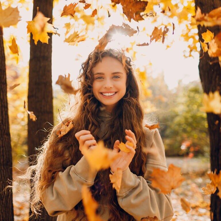 Herbstbild einer jungen Frau, die im Wald und im Sonnenschein in die Kamera lächelt. Um sie herum fallen Herbstblätter auf den Boden. Symbolbild für die vielen Gründe, warum der November eingentlich ein ganz toller Monat ist.  (Foto: Adobe Stock, Adobe Stock | primipil)