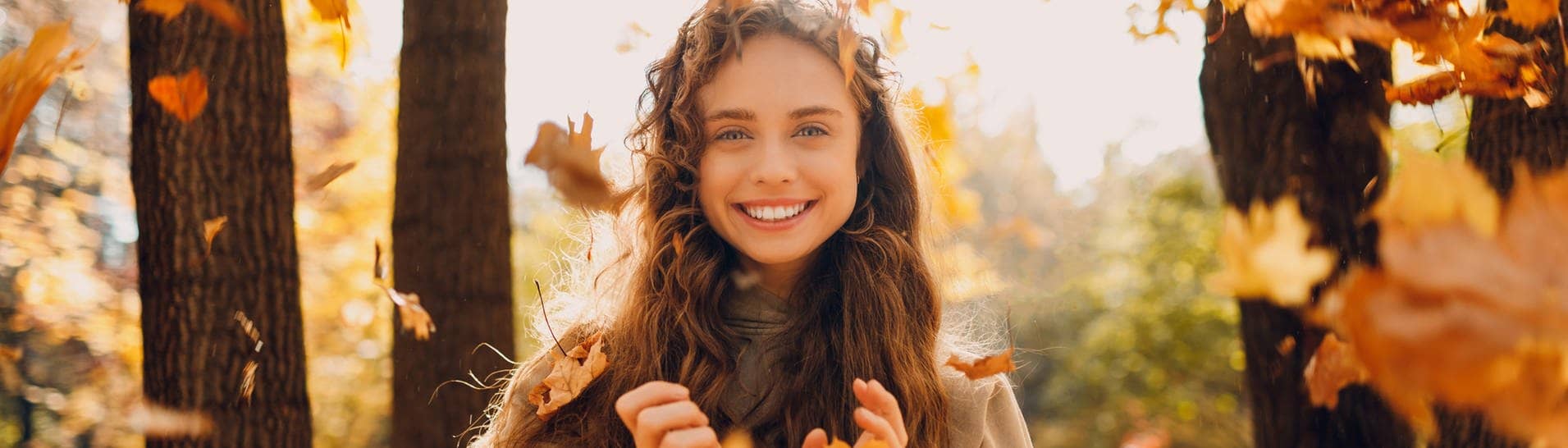 Herbstbild einer jungen Frau, die im Wald und im Sonnenschein in die Kamera lächelt. Um sie herum fallen Herbstblätter auf den Boden. Symbolbild für die vielen Gründe, warum der November eingentlich ein ganz toller Monat ist.  (Foto: Adobe Stock, Adobe Stock | primipil)