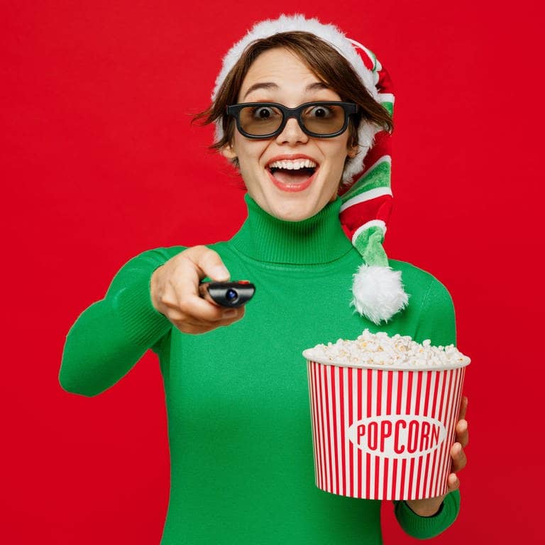 Symbolbild für das Schauen von Weihnachtsfilmen: Junge aufgeregte Frau mit 3D-Brille trägt grünen Rollkragenpullover mit Weihnachtsmannmütze, schaut sich einen Film an und hält einen entfernten Popcorn-Eimer im Kino, isoliert auf schlichtem rotem Hintergrund. (Foto: Adobe Stock, Adobe Stock | ViDi Studio)