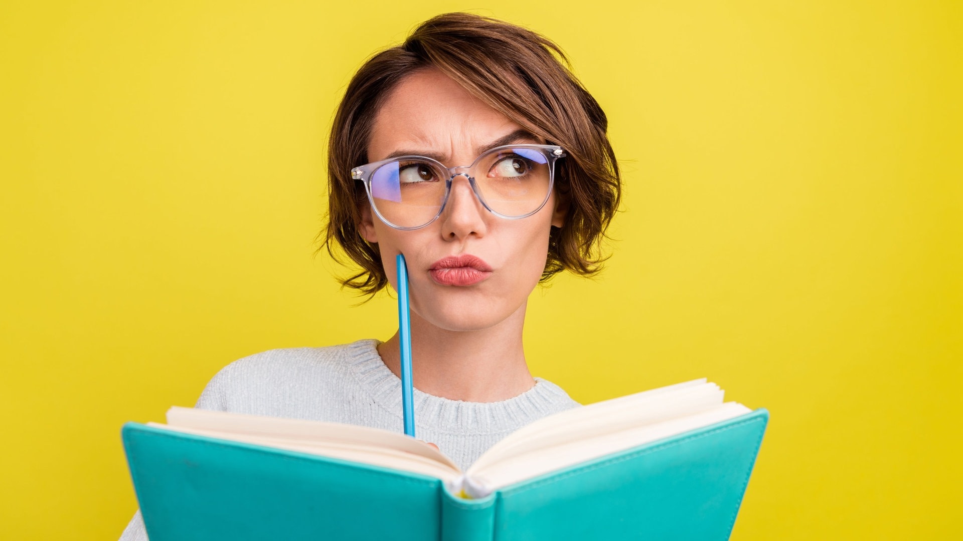 Foto: Adobe Stock, deagreez Junge Frau denkt über verrückte Schreibweisen nach, die vom Duden anerkannt wird. Sie schaut denkend in die Luft, hält ein Buch in der Hand und einen Stift an die Wange. (Foto: Adobe Stock, deagreez)