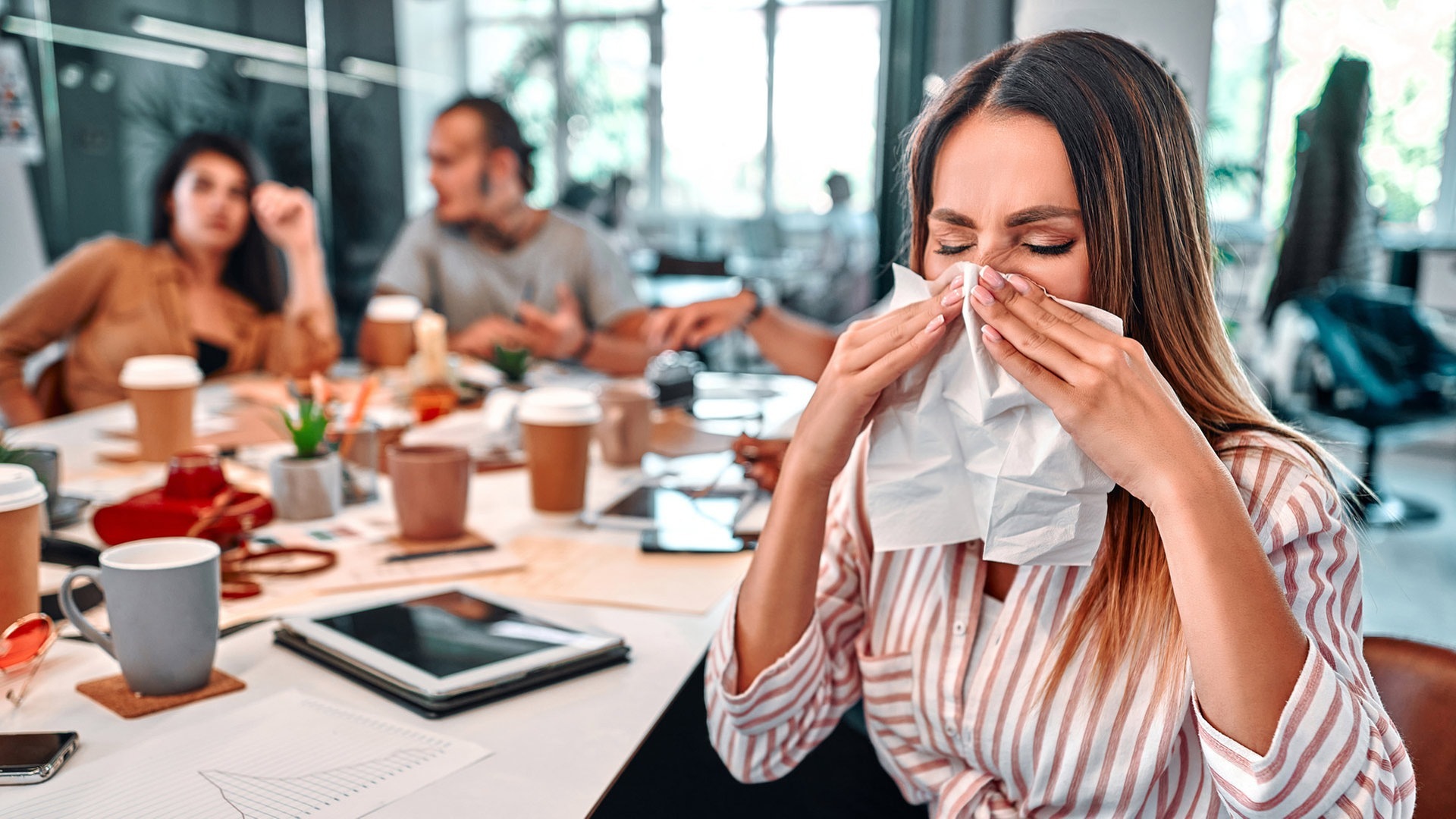 Junge Frau sitzt im Büro, hinter ihr Arbeitskollegen. Sie schnäuzt krank die Nase, vielleicht hat sie Corona, auf jeden Fall geht sie krank arbeiten. (Foto: Adobe Stock, HBS)