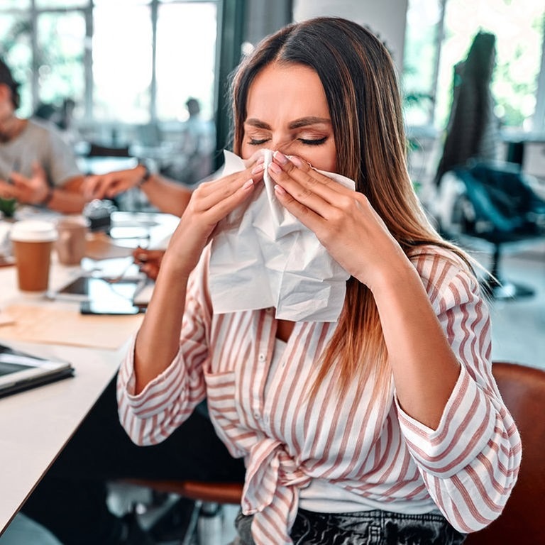 Junge Frau sitzt im Büro, hinter ihr Arbeitskollegen. Sie schnäuzt krank die Nase, vielleicht hat sie Corona, auf jeden Fall geht sie krank arbeiten. (Foto: Adobe Stock, HBS)