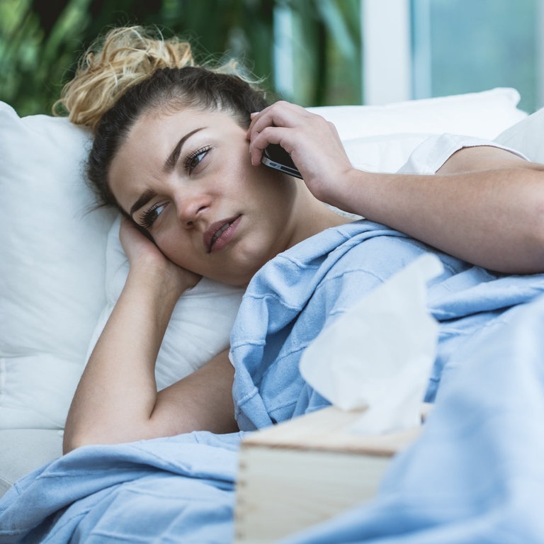 Die telefonische Krankmeldung beim Arbeitgeber macht es für so manchen Arbeitnehmer einfacher: Hier liegt eine Frau im Bett mit Telefon am Ohr und Taschentüchern auf dem Bett. (Foto: IMAGO, Pond5 Images)