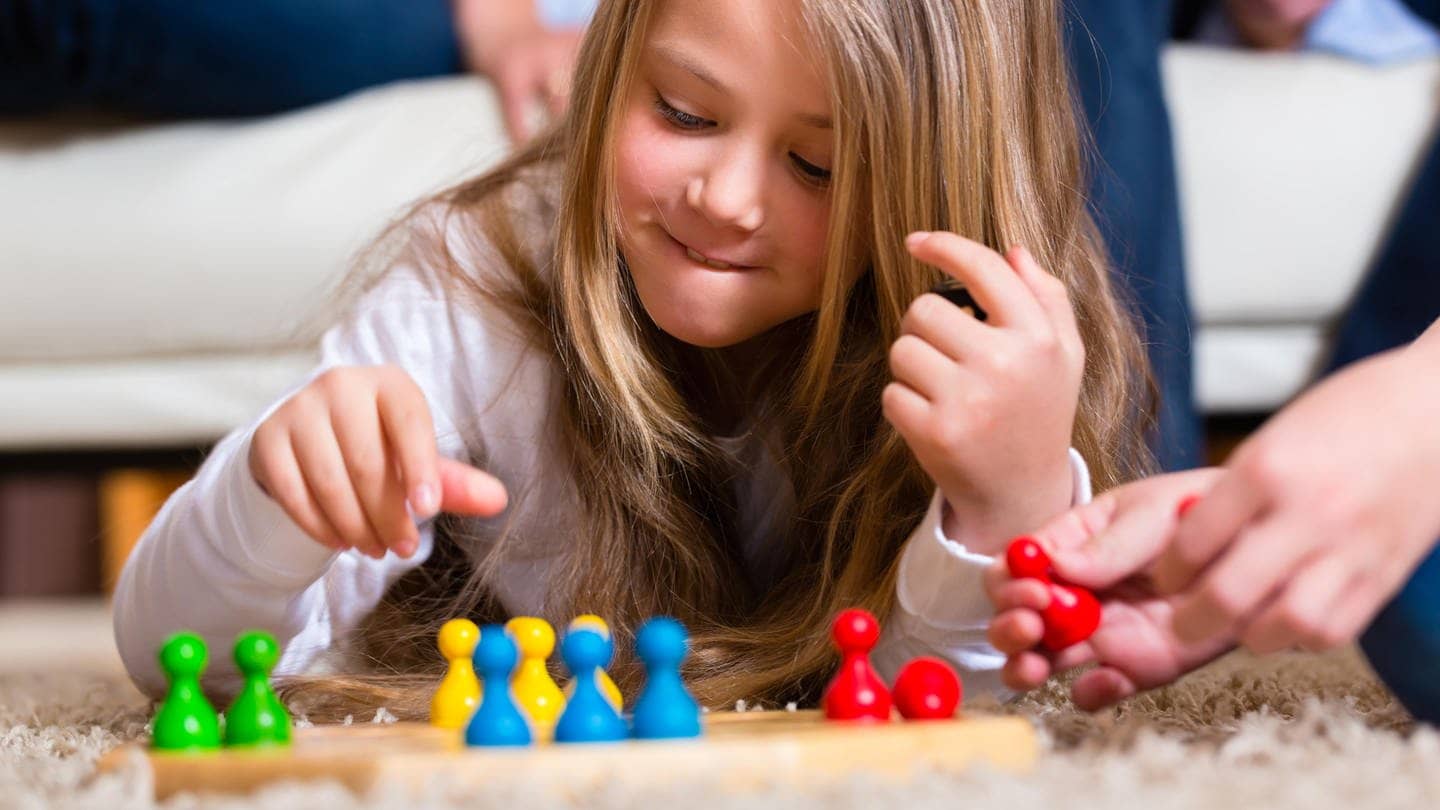 Kleines Mädchen spielt „Mensch ärgere dich nicht“, vor ihr ist das Spielfeld und in der Hand hält sie eine Spielfigur. (Foto: Adobe Stock, Kzenon)