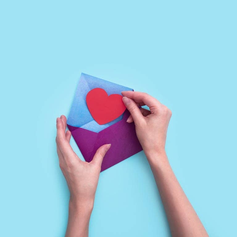 Eine Hand steckt ein rotes Papierherz in einen lila Umschlag. (Foto: Adobe Stock, Vladimir Zlotnik)