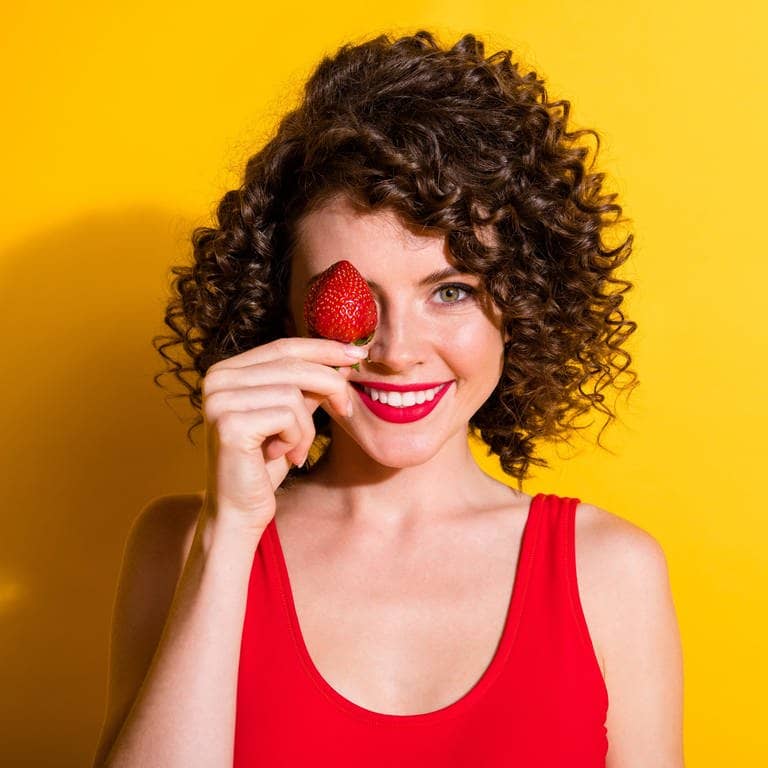 Junge Frau mit braunen Locken hält sich eine rote Erdbeere vors Gesicht und lacht. Symbolbild für Wissen und Fakten rund um Erdbeeren. (Foto: Adobe Stock, deagreez)