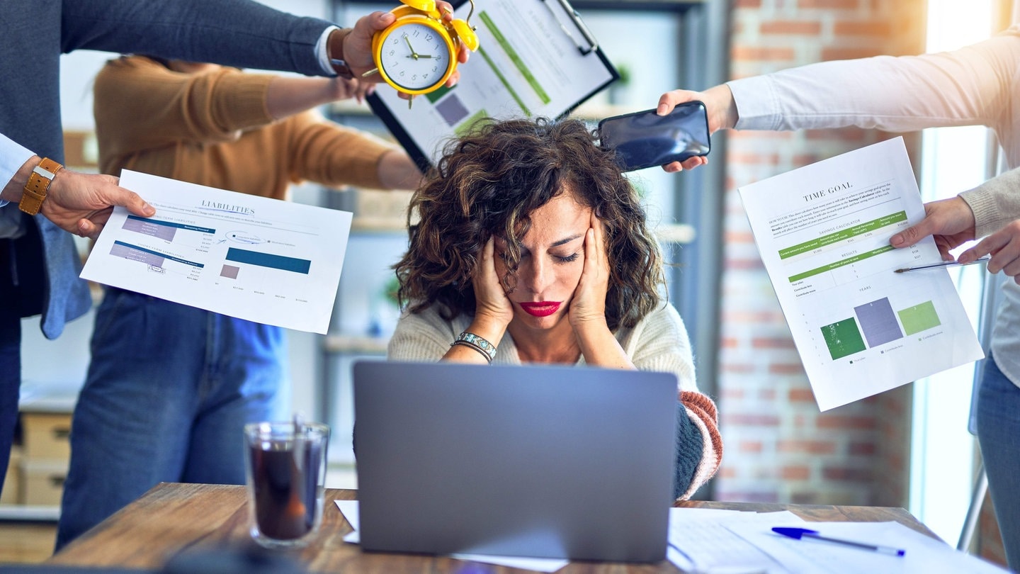 Krakenimages.com (Foto: Adobe Stock, Junge Frau stützt überfordert den Kopf in die Hände, sie sitzt im Büro vor einem Laptop, um sie herum strecken ihr Menschen Arbeitsutensilien entgegen. Symbolbild für Überstunden und Mehrarbeit im Job, die überfordert.)