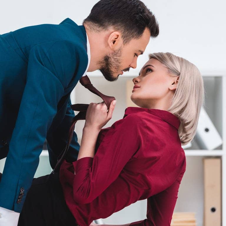 Mann und Frau im Business-Look liegen verführerisch auf dem Schreibtisch, sie zieht ihn an der Krawatte zu sich (Foto: Adobe Stock/LIGHTFIELD STUDIOS)