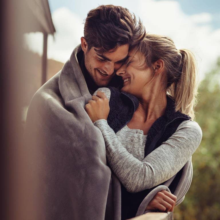 Junges Paar kuschelt verliebt in einer Decke und lächelt sich gegenseitig an