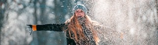 Junge Frau, warm angezogen mit Mütze, wirft Schnee in die Luft und freut sich