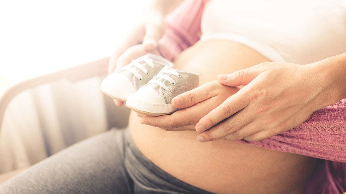 Eine Frau mit Babybauch, die zusammen mit ihrem Partner kleine Babyschühchen darauf hält.