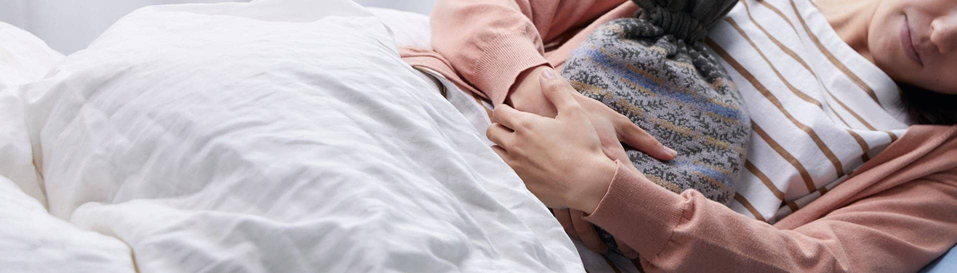 Eine Frau liegt im Bett und hält eine Wärmflasche an ihren Bauch, um ihre Regelschmerzen zu lindern (Foto: Imago)