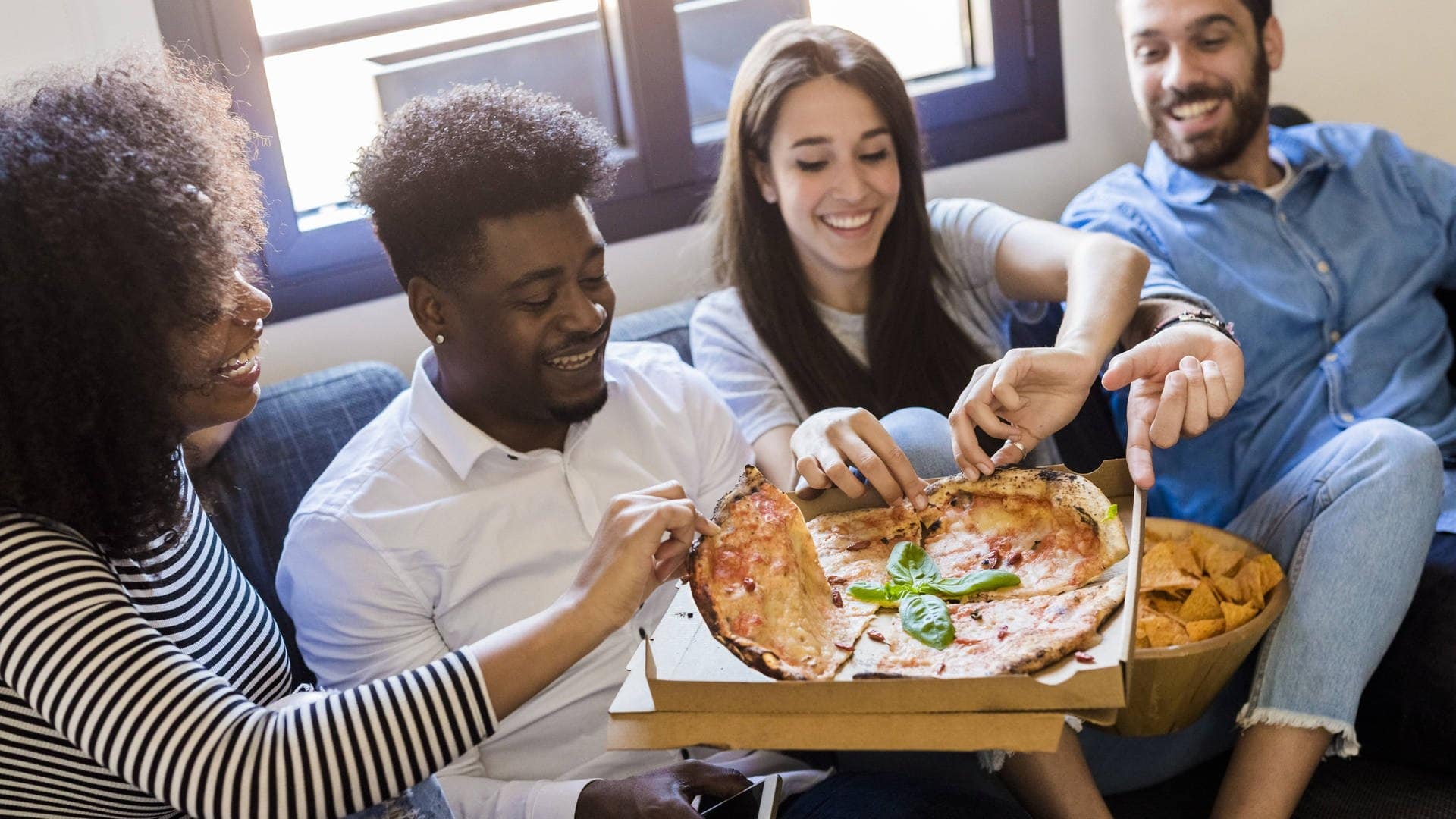 4 Leute, möglicherweise Partner, sitzen auf einem Sofa in einer Wohnung nach dem Einziehen und teilen sich eine Pizza, die in der Mitte gehalten wird.