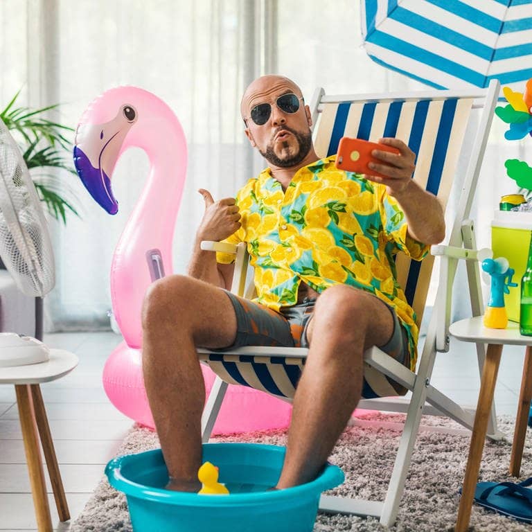 Junger Mann mit Sonnenbrille und buntem Hemd sitzt auf einem Liegestuhl zu Hause, das aussieht wie im Urlaub – neben ihm ein Ventilator und ein aufblasbarer Flamingo und ein Sonnenschirm. Er zeigt mit dem Daumen nach oben und hält ein Handy für das Urlaubsfoto vors Gesicht.