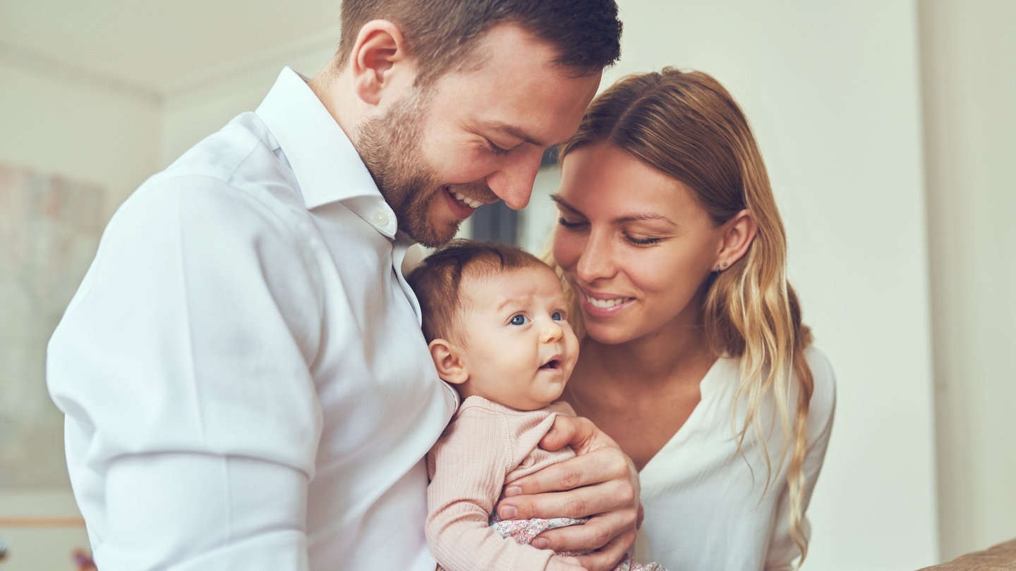 Junge Eltern mit ihrem Baby. Der Vater hält lachend das Baby auf dem Arm, die Mutter lächelt das Kind ebenfalls an.