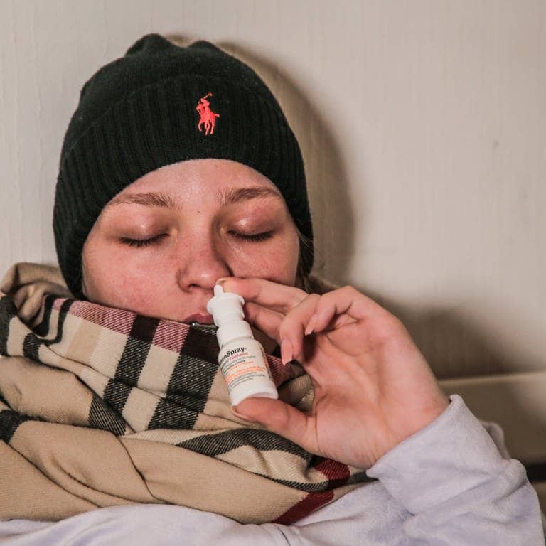Kranke Frau mit Schal und schwarzer Mütze nutzt ein Nasenspray (Foto: IMAGO, impfung-gegen-grippe)