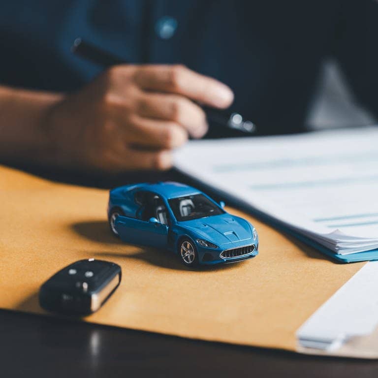 Blaues Miniaturauto liegt auf einem Tisch. Daneben ein richtiger Autoschlüssel, im Hintergrund eine hand mit einem Stift.