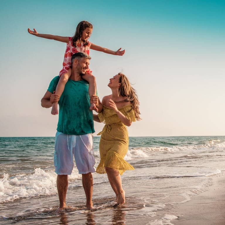 Eine junge Familie spaziert am Strand. Mann, Frau und ein Kind auf den Schultern des Vaters im Urlaub. Es drohen keine Konsequenzen und es ist keine Arbeitsverweigerung, wenn sie sich von hier aus nicht zurück zur Arbeit holen lassen.