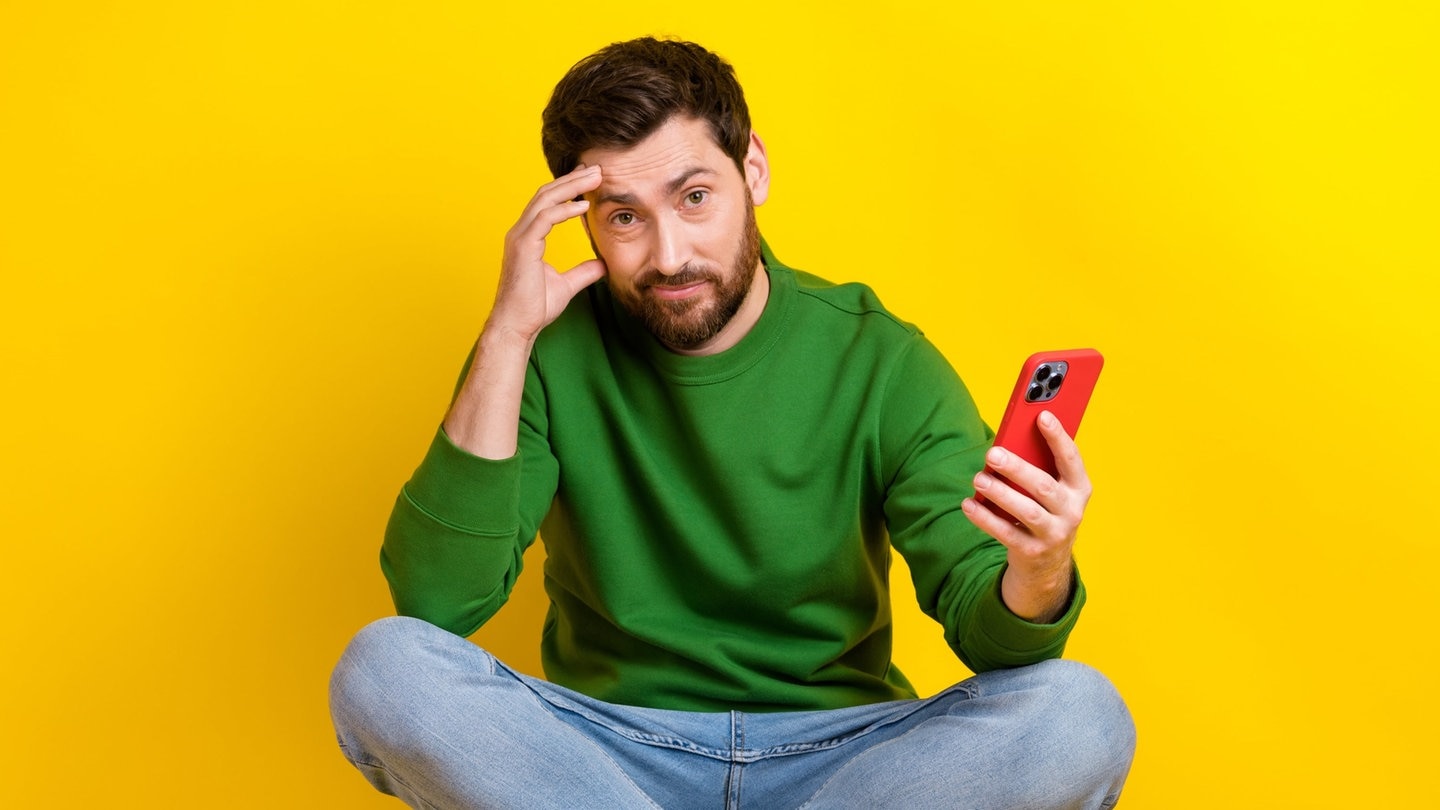 Junger Mann sitzt im Schneidersitz und mit dem Handy in der Hand auf einem Hocker. Er häkt denkend die Hand an den Kopf. Auf seinem Smartphone sieht er bestimmt gerade die überraschenden alternativen Schreibweisen laut Duden und wundert sich.