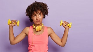 Junge Frau im Sport-Outfit und mit Hanteln in der Hand. Sie sieht genervt und wenig motiviert aus, hat wohl keine Lust oder ist ein Fitnessmuffel.
