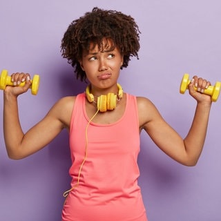 Junge Frau im Sport-Outfit und mit Hanteln in der Hand. Sie sieht genervt und wenig motiviert aus, hat wohl keine Lust oder ist ein Fitnessmuffel.