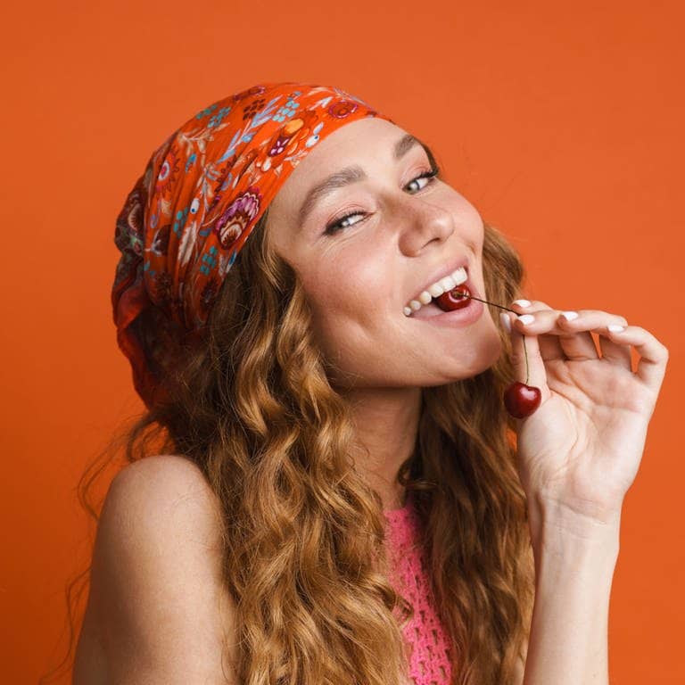 Junge Frau mit buntem Tuch in den Haaren beißt an einer Kirsche – getreu dem Motto „with me is not good cherry eating“, einer deutschen Redewendung, wörtlich übersetzt auf Englisch.