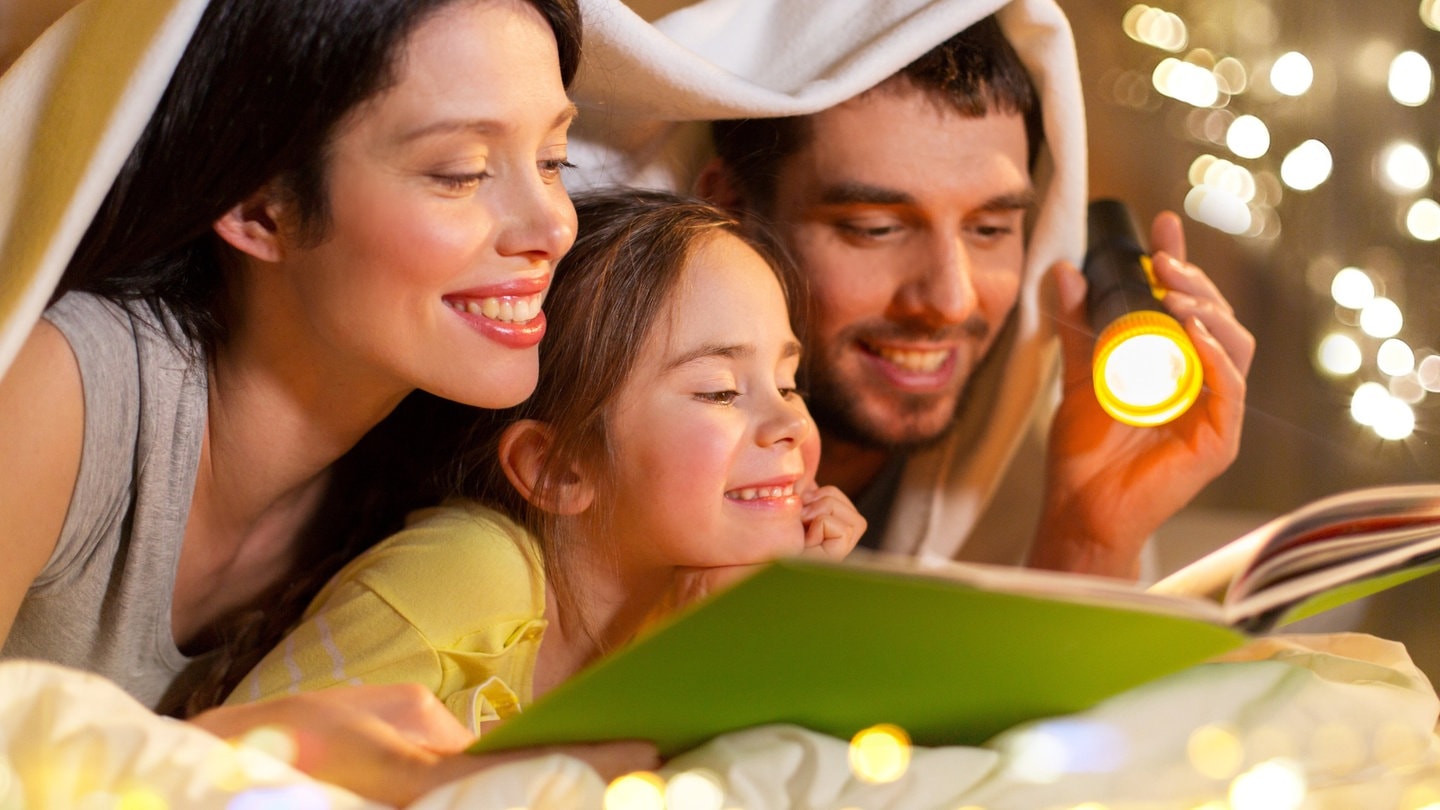 Eine Familie mit einem Kind liegt auf dem Boden mit einem Buch vor sich und hält eine Taschenlampe in der Hand (Foto: picture-alliance / Reportdienste, picture alliance / Zoonar | lev dolgachov)