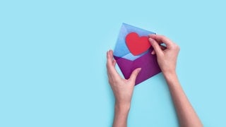 Eine Hand steckt ein rotes Papierherz in einen lila Umschlag.