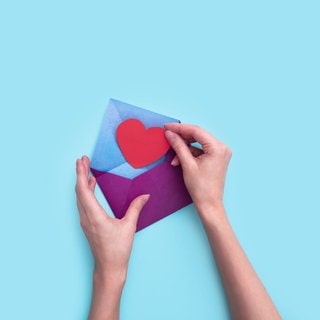 Eine Hand steckt ein rotes Papierherz in einen lila Umschlag.