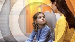 Mutter sitzt mit Tochter auf dem Sofa, Kind schaut Mutter erwartungsvoll an.