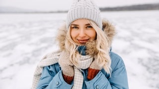 Junge Frau in Winter-Kleidung mit Mütze und Mantel steht in einer Schneelandschaft in der Kälte. Vielleicht denkt sie über Mythen rund um die kalte Jahreszeit nach.