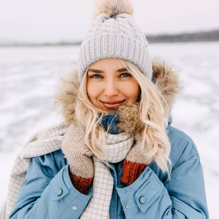 Junge Frau in Winter-Kleidung mit Mütze und Mantel steht in einer Schneelandschaft in der Kälte. Vielleicht denkt sie über Mythen rund um die kalte Jahreszeit nach.
