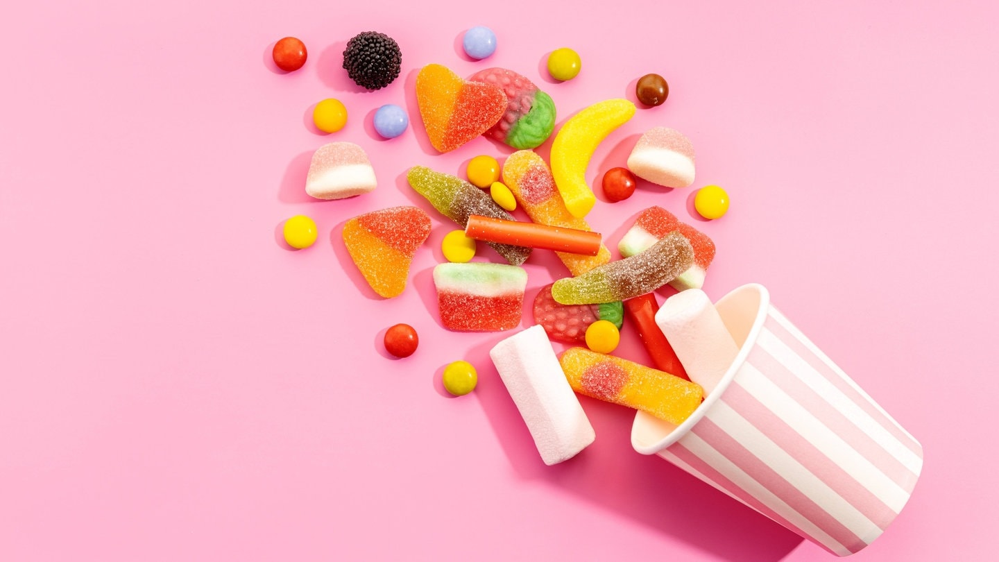 Bunte Fruchtgummis fallen aus einem gestreiften Papierbecher (Foto: IMAGO, Addictive Stock)